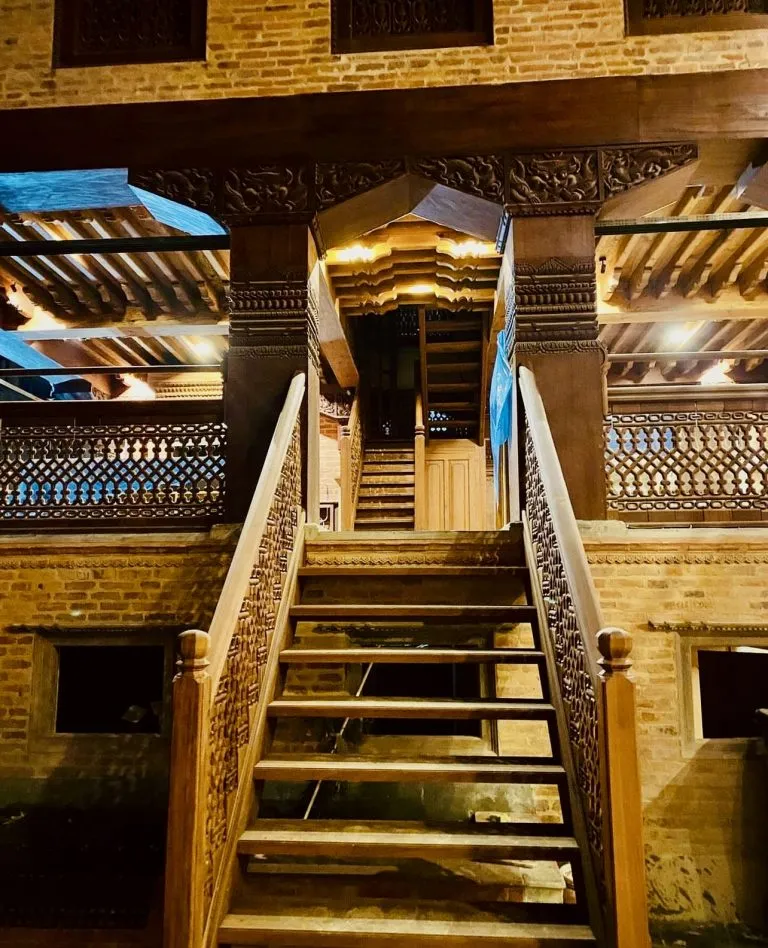 Carved Newari wooden staircase rising into the lit courtyard of the restored heritage building at night.
