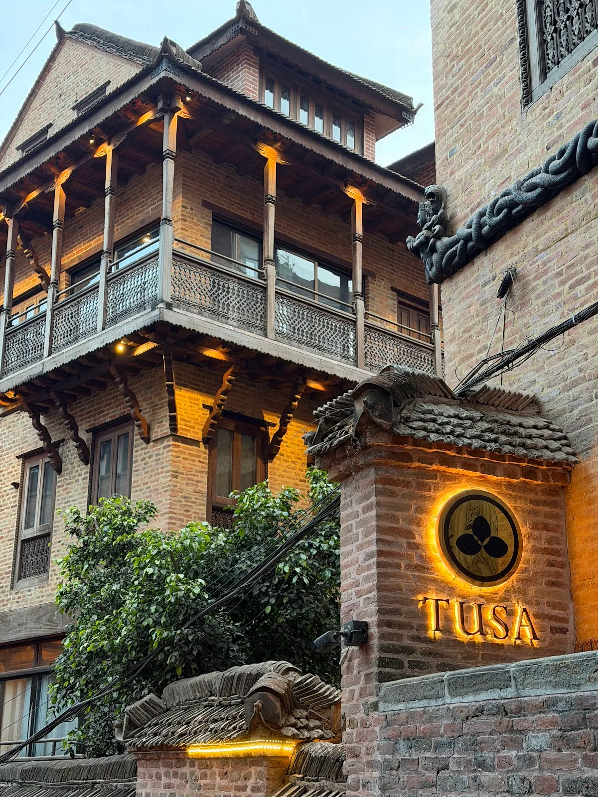 The lit facade of Restaurant Tusa, a restored Malla-era brick courtyard in Bhaktapur, at dusk with the Tusa monogram glowing on the gate.